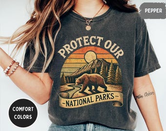 Puede incluir: Una camiseta gris oscuro con un gráfico de estilo vintage de un oso en un bosque con el texto "Protege nuestros parques nacionales".