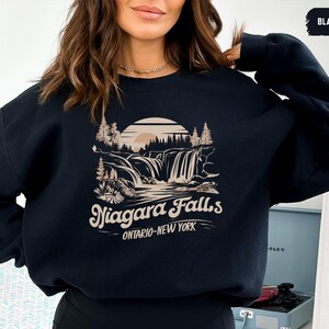 Puede incluir: Sudadera negra con un gráfico de las Cataratas del Niágara en beige y blanco. El diseño incluye una escena de cascada con árboles, una puesta de sol y el texto "Niagara Falls Ontario-New York."