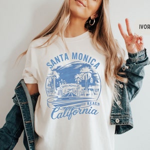 Può includere: Una maglietta bianca con un design grafico blu che dice "Santa Monica Beach California". La grafica presenta un'illustrazione stilizzata del molo di Santa Monica con palme e le parole "Santa Monica" in uno stendardo.
