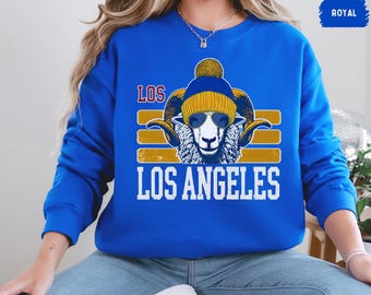 May include: A royal blue sweatshirt featuring a graphic of a ram wearing a blue and yellow beanie and sunglasses. The text "LOS ANGELES" is printed below the ram.
