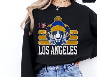 May include: Black sweatshirt with a graphic of a ram wearing a blue and yellow beanie and sunglasses. The graphic is surrounded by a yellow, orange, and white striped design. The text "LOS ANGELES" is printed below the graphic.