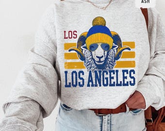 May include: A gray sweatshirt with a graphic of a ram wearing a blue and yellow beanie and sunglasses. The graphic says "Los Angeles" with the word "Los" in blue and "Angeles" in yellow. The sweatshirt has a distressed look and is made of a soft, comfortable material.