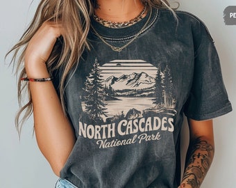 Może przedstawiać: Szary t-shirt z nadrukiem w stylu vintage przedstawiającym pasmo górskie i jezioro z tekstem "North Cascades National Park".