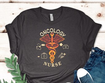 May include: Dark gray t-shirt with the words "ONCOLOGY NURSE" and a medical symbol design in orange and yellow. The shirt is paired with blue jeans and navy blue sneakers with white soles. A sprig of green leaves is in the corner.