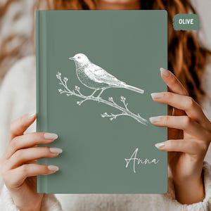 Peut inclure: Journal vert olive avec une illustration blanche d'un oiseau sur une branche. Le nom "Anna" est écrit en cursive en bas. Le journal est de forme rectangulaire.