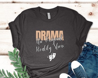 May include: A dark gray t-shirt with the text "DRAMA the original Reality Show" in a light brown color. Two white theater masks are below the text.