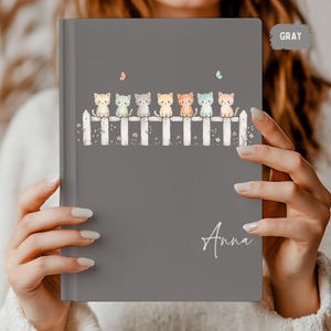 Könnte beinhalten: Graues Notizbuch mit einer Illustration von bunten Kätzchen auf einem weißen Zaun. Der Name "Anna" ist kursiv geschrieben. Das Wort "GRAY" befindet sich in einem grauen Rechteck oben rechts.