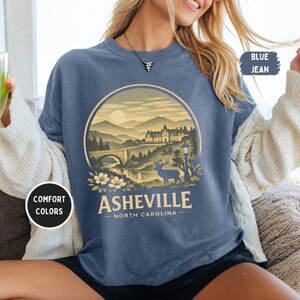 May include: Blue jean-colored t-shirt with a circular graphic featuring a mountain landscape, bridge, and deer. The text "Asheville North Carolina" is printed below the graphic. The shirt also has the text "Comfort Colors" on a white circle.