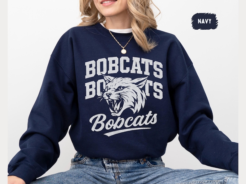 Puede incluir: Sudadera azul marino con la palabra "BOBCATS" en letras blancas y un gr&aacute;fico de lince blanco. La sudadera tiene cuello redondo y mangas largas. La palabra "BOBCATS" tambi&eacute;n est&aacute; escrita en una fuente de escritura.