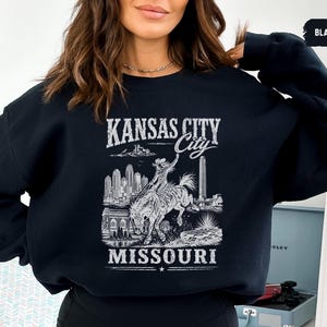 May include: Black sweatshirt with a white graphic featuring the text "KANSAS CITY MISSOURI". The graphic includes an illustration of a cowboy riding a horse, a cityscape, and other architectural elements.