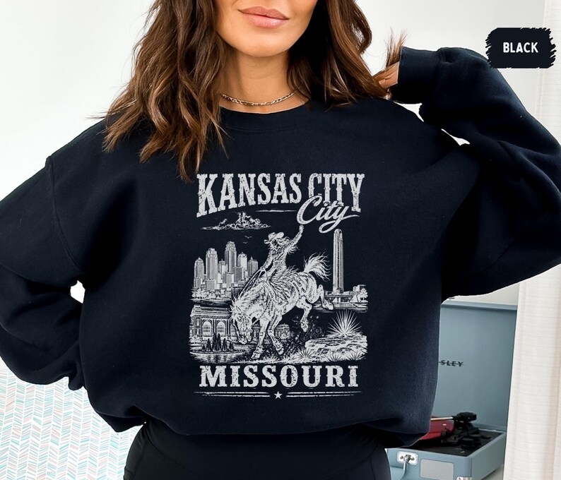 May include: Black sweatshirt with a white graphic featuring the text "KANSAS CITY MISSOURI". The graphic includes an illustration of a cowboy riding a horse, a cityscape, and other architectural elements.