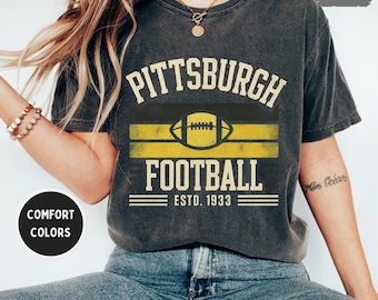 May include: A charcoal grey t-shirt featuring the text "PITTSBURGH FOOTBALL" in a vintage font, with a football graphic and the text "ESTD. 1933". The shirt has yellow and black stripes, and the words "COMFORT COLORS" are on a black circle.