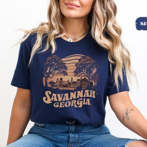 Puede incluir: Una camiseta azul marino con un gr&aacute;fico de estilo vintage del horizonte de Savannah, Georgia. El gr&aacute;fico presenta una puesta de sol, &aacute;rboles y edificios. El texto "Savannah Georgia" est&aacute; impreso en marr&oacute;n debajo del gr&aacute;fico.