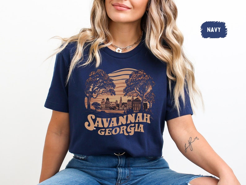 Puede incluir: Una camiseta azul marino con un gr&aacute;fico de estilo vintage del horizonte de Savannah, Georgia. El gr&aacute;fico presenta una puesta de sol, &aacute;rboles y edificios. El texto "Savannah Georgia" est&aacute; impreso en marr&oacute;n debajo del gr&aacute;fico.