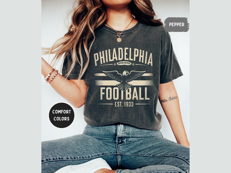 May include: A dark grey t-shirt with a vintage-style graphic design featuring the text "Philadelphia Football Est. 1933" and an eagle graphic. The shirt has a distressed look and is made of a soft, comfortable fabric.