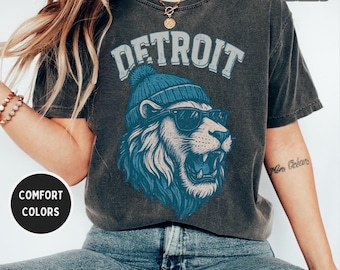 Op de afbeelding: Donkergrijs t-shirt met het woord "DETROIT" in lichtblauwe letters boven een leeuwengrafiek. De leeuw draagt een blauwe muts en zonnebril. Het shirt is een comfort kleur.