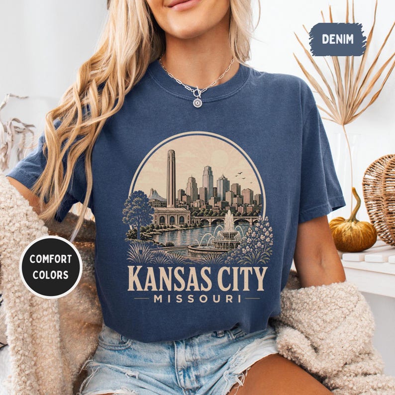 May include: Denim blue t-shirt featuring a circular graphic of the Kansas City skyline, including a tall tower, buildings, and a fountain. The text "Kansas City Missouri" is printed below the graphic. The shirt has "Comfort Colors" and "Denim" tags.