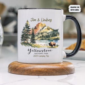 Op de afbeelding: Een witte keramische mok met een zwarte handgreep. De mok heeft een aquarel-illustratie van een berglandschap met een meer en een bizon. De tekst "Jim & Lindsey" staat boven de afbeelding, en de tekst "Yellowstone National Park 2025 Camping Trip" staat onder de afbeelding.