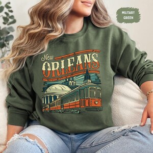 May include: A green sweatshirt with a vintage-style graphic of a streetcar in New Orleans. The text "New Orleans" is printed in a bold, retro font.