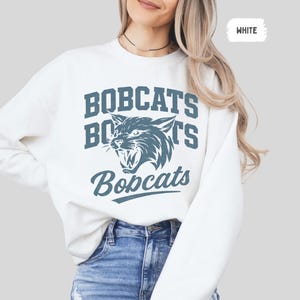 Puede incluir: Sudadera blanca con la palabra "BOBCATS" en letras azules y un gr&aacute;fico azul de un lince pardo gru&ntilde;endo. La palabra "Bobcats" tambi&eacute;n est&aacute; impresa en una fuente cursiva debajo del gr&aacute;fico. La palabra "WHITE" est&aacute; en un rect&aacute;ngulo blanco en la esquina superior derecha.