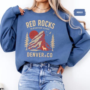 May include: Indigo blue sweatshirt featuring a graphic of Red Rocks in Denver, Colorado. The design includes a red sun, a deer, and the text "RED ROCKS ELEVATION 6,450' DENVER CO".