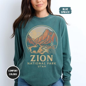 Pode incluir: Camisola de manga comprida azul abeto com um gráfico circular do Parque Nacional de Zion. O gráfico inclui um carneiro, um rio e montanhas em tons de castanho, laranja e verde. O texto "ZION NATIONAL PARK UTAH" está abaixo.
