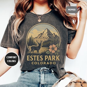 May include: Dark gray t-shirt with a circular graphic featuring a mountain range, a deer, and a flower. The text "Estes Park Colorado" is printed below the graphic. The shirt is labeled "Comfort Colors" and the color is "Pepper".