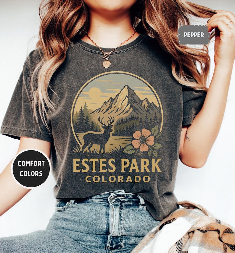 May include: Dark gray t-shirt with a circular graphic featuring a mountain range, a deer, and a flower. The text "Estes Park Colorado" is printed below the graphic. The shirt is labeled "Comfort Colors" and the color is "Pepper".