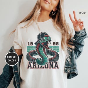 May include: White t-shirt with a green snake wearing a black baseball cap and sunglasses. The snake is surrounded by a black and white striped design with the text "19 98 ARIZONA".