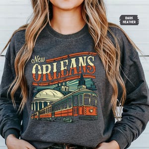 May include: Dark heather grey sweatshirt with a vintage-style graphic of a streetcar in New Orleans. The graphic includes the text "New Orleans" in a retro font.