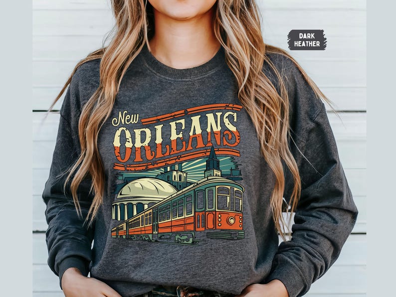May include: Dark heather grey sweatshirt with a vintage-style graphic of a streetcar in New Orleans. The graphic includes the text "New Orleans" in a retro font.