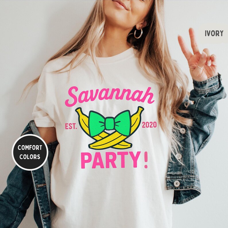 May include: Ivory-colored t-shirt with a graphic design. The design features the name "Savannah" in pink, a green bow, two yellow bananas, and the word "PARTY!" in pink. The text "EST. 2020" is also included.