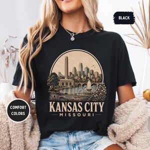 May include: Black t-shirt featuring a circular graphic of the Kansas City skyline, with the text "Kansas City Missouri". The design includes a river, fountain, and buildings. The shirt has "Comfort Colors" and "Black" labels.