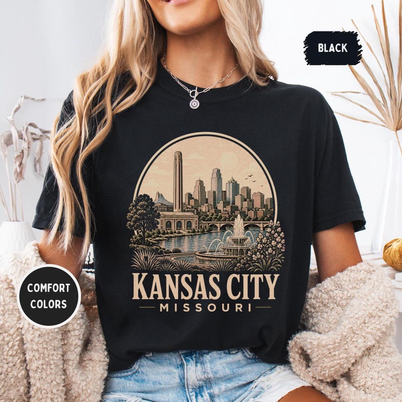 May include: Black t-shirt featuring a circular graphic of the Kansas City skyline, with the text "Kansas City Missouri". The design includes a river, fountain, and buildings. The shirt has "Comfort Colors" and "Black" labels.