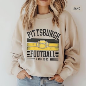 May include: Sand-colored sweatshirt featuring a Pittsburgh Football design. The design includes the word "PITTSBURGH" above a football graphic and the word "FOOTBALL" with "ESTD. 1933" below. The design is in black and yellow.