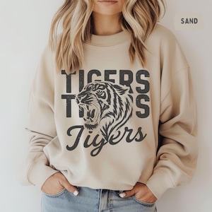 May include: Sand-colored sweatshirt with the word "Tigers" in a bold, distressed font, with a roaring tiger graphic. The word "Tigers" is repeated in a stacked design. The sweatshirt is a neutral color and has a classic crew neck.