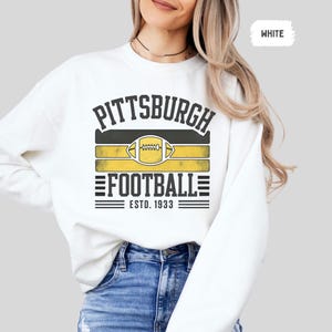 May include: White sweatshirt featuring a Pittsburgh Football design. The design includes the word "Pittsburgh" above a football graphic and the word "Football" with "ESTD. 1933" below. The football graphic is yellow and black.