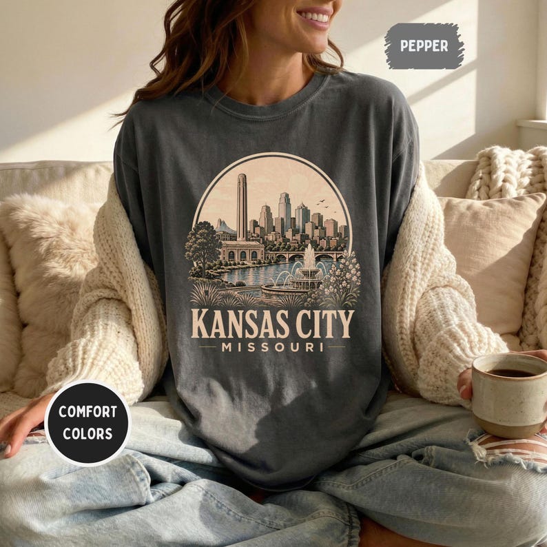 May include: Dark gray t-shirt with a circular graphic featuring a Kansas City skyline, a lake, and the text "Kansas City Missouri." The shirt is part of the Comfort Colors collection.