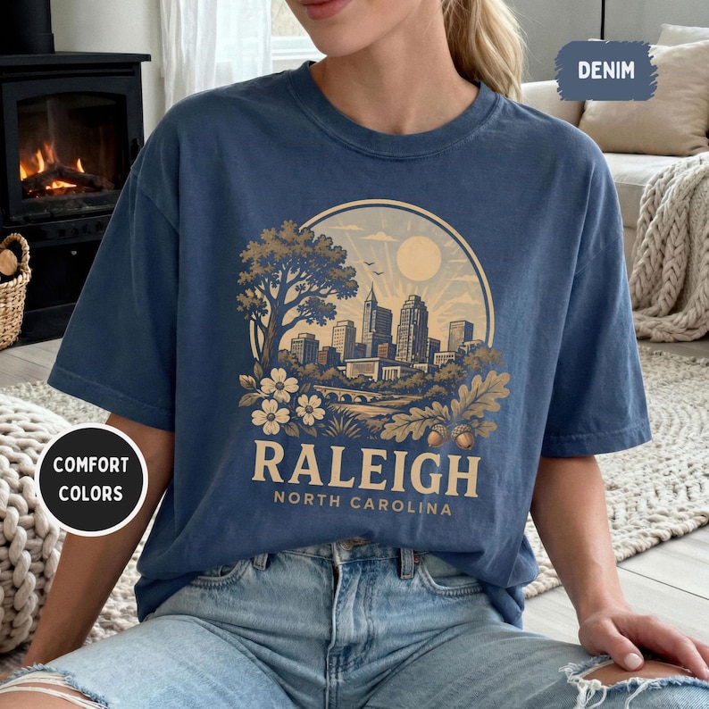 May include: A denim blue t-shirt featuring a circular graphic of the Raleigh, North Carolina skyline, with the text "RALEIGH NORTH CAROLINA". The design includes a tree, flowers, and a bridge. The shirt has short sleeves.