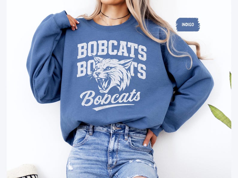 May include: Indigo blue sweatshirt with the word "BOBCATS" in white block letters above a white graphic of a snarling wildcat. The word "Bobcats" is also written in a script font below the graphic. The sweatshirt has long sleeves and a crew neck.