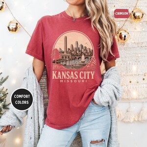 May include: Crimson-colored t-shirt featuring a circular graphic of the Kansas City skyline, with the text "Kansas City Missouri" below. The shirt has a relaxed fit and is part of the Comfort Colors collection.