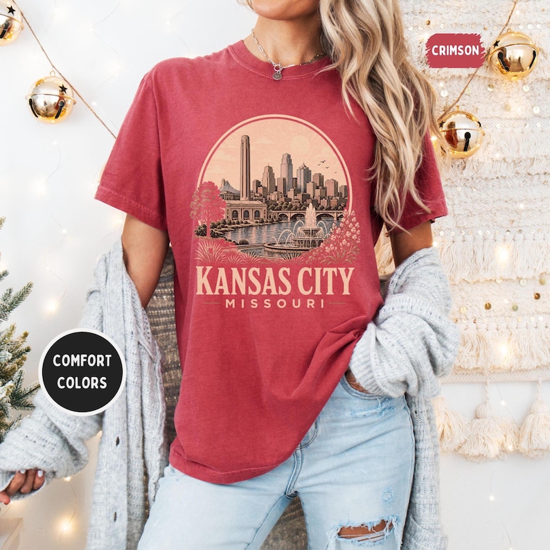 May include: Crimson-colored t-shirt featuring a circular graphic of the Kansas City skyline, with the text "Kansas City Missouri" below. The shirt has a relaxed fit and is part of the Comfort Colors collection.
