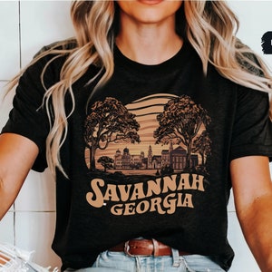 Puede incluir: Una camiseta negra con un dise&ntilde;o gr&aacute;fico marr&oacute;n de una ciudad con &aacute;rboles y el texto "Savannah Georgia".