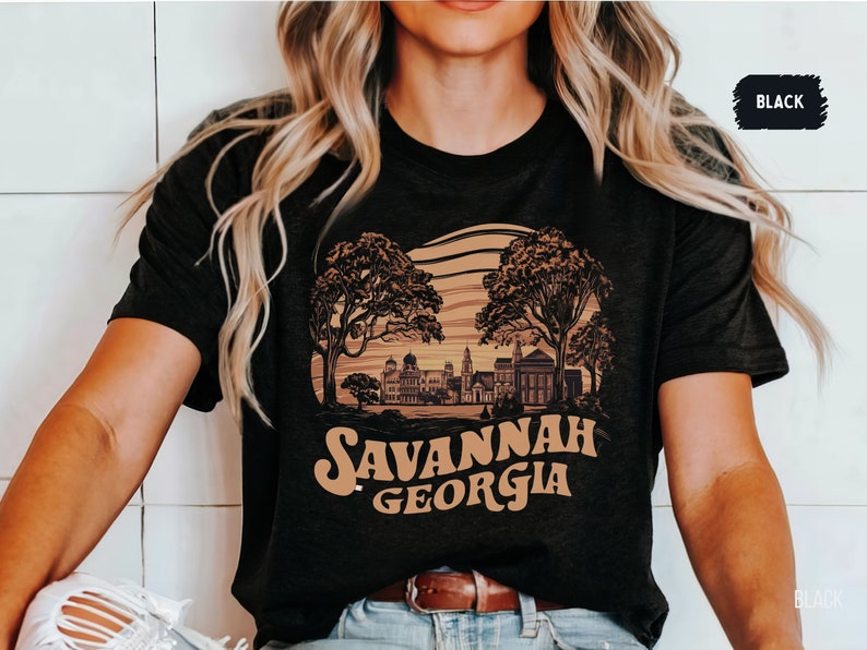 Puede incluir: Una camiseta negra con un dise&ntilde;o gr&aacute;fico marr&oacute;n de una ciudad con &aacute;rboles y el texto "Savannah Georgia".