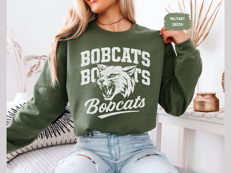 Puede incluir: Sudadera verde militar con la palabra "BOBCATS" en letras blancas en bloque encima y debajo de un gr&aacute;fico blanco de un lince rugiente. La sudadera es de manga larga y cuello redondo.