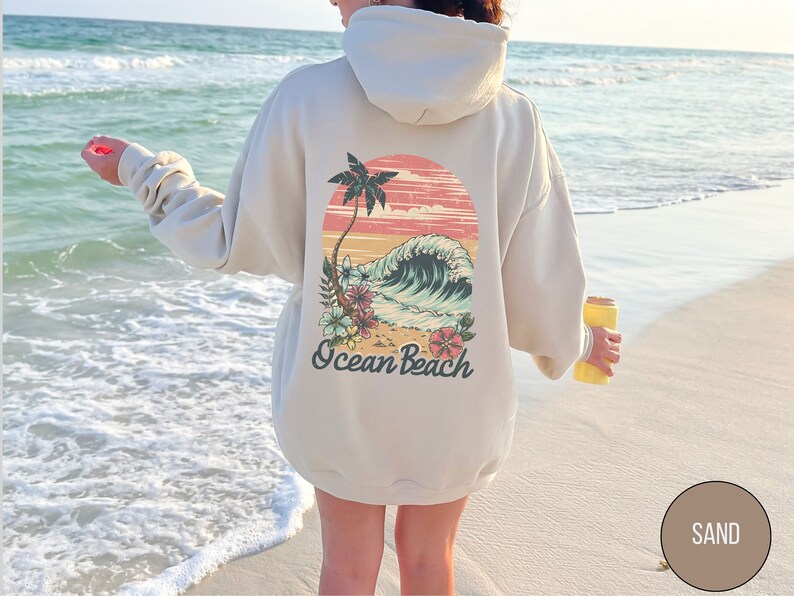 Op de afbeelding: Beige hoodie met een retro-afbeelding op de rug met een palmboom, oceaangolf en bloemen. De tekst "Ocean Beach" staat eronder. De hoodie wordt gedragen op een zandstrand.