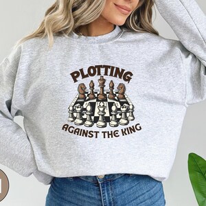 May include: A gray sweatshirt with the text "Plotting Against The King" and a graphic of a chessboard with chess pieces.