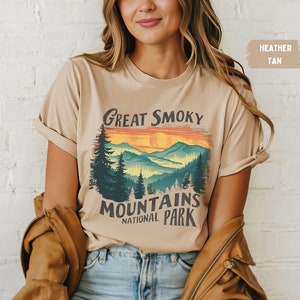 May include: A heather tan t-shirt with a vintage-style graphic of the Great Smoky Mountains National Park. The graphic features a sunset over the mountains with a silhouette of trees in the foreground.