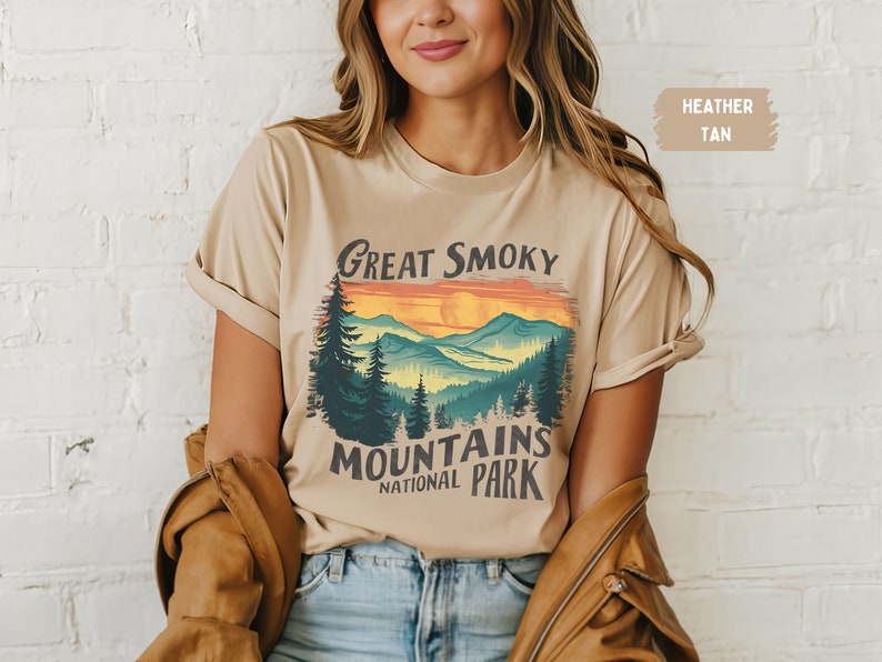 May include: A heather tan t-shirt with a vintage-style graphic of the Great Smoky Mountains National Park. The graphic features a sunset over the mountains with a silhouette of trees in the foreground.