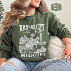 May include: Military green sweatshirt with a white graphic of Kansas City, Missouri. The design features a cowboy riding a horse, city skyline, and the text "Kansas City Missouri". The sweatshirt is a crew neck style.